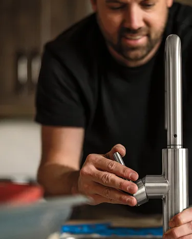 Plumber/installer in kitchen with dark cabinets (background) installing chrome single-handle Delta faucet (foreground).